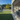 An outdoor patio area with a large green artificial turf lawn, gray composite deck flooring, a covered seating area with white columns, and a backyard pool and trees in the distance under a clear blue sky.