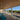 A spacious covered porch with a wood ceiling and composite floor, white railings, and bright blue Adirondack chairs. Beyond the porch, there is a scenic view of green dunes, a boardwalk, and the ocean under a clear blue sky.