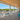 A spacious covered porch with gray composite decking, white railings, a wood ceiling, and bright turquoise Adirondack chairs. Large windows and glass doors offer a view of green dunes and blue sky, with a pool visible below.